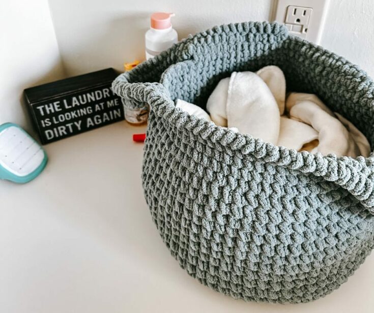 A textured gray laundry basket filled with white clothes sits on a counter near assorted items and a sign that reads, The laundry is looking at me dirty again.