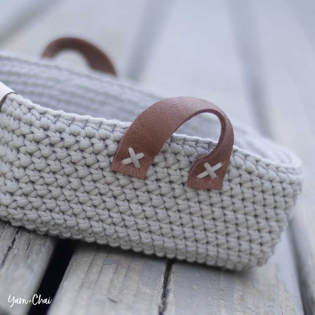 Close-up of a crocheted basket with two brown leather handles, each attached with white stitched Xs, placed on a wooden surface.