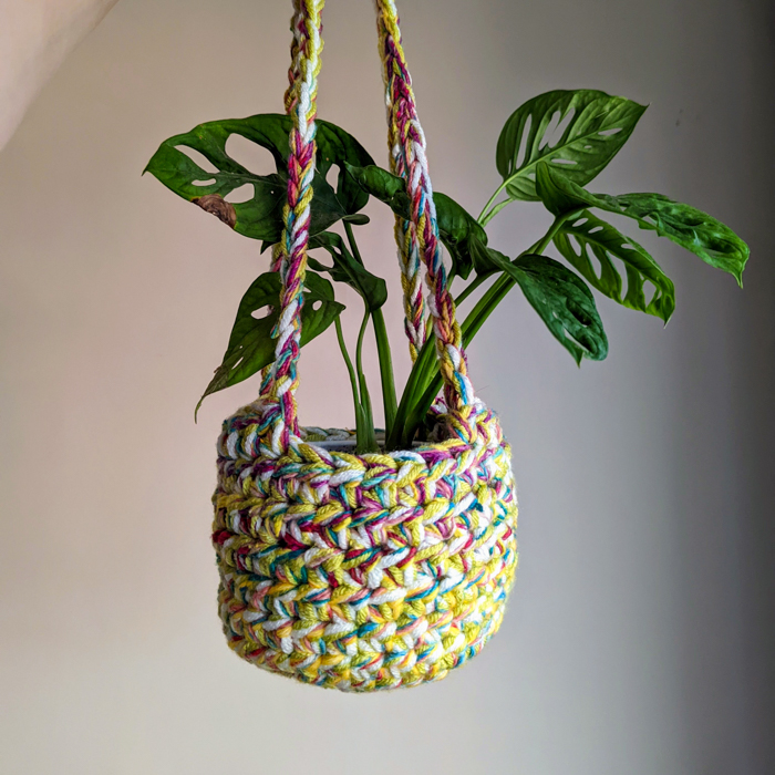A green potted plant in a colorful, hand-crocheted hanging basket against a plain background.