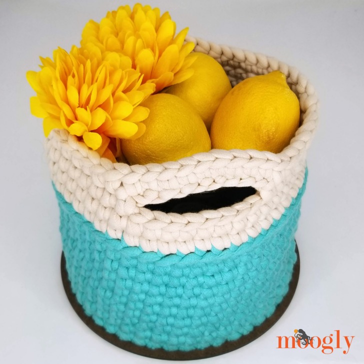 A crocheted basket with blue and cream sides holds three lemons and two yellow artificial flowers; the basket has a handle slot and sits on a round base.