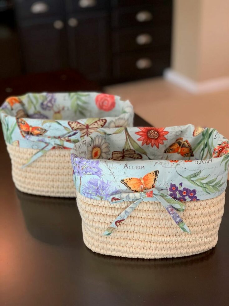 Two woven baskets with floral and butterfly-patterned fabric liners sit on a dark surface. Each basket has a bow tied from the same patterned fabric.