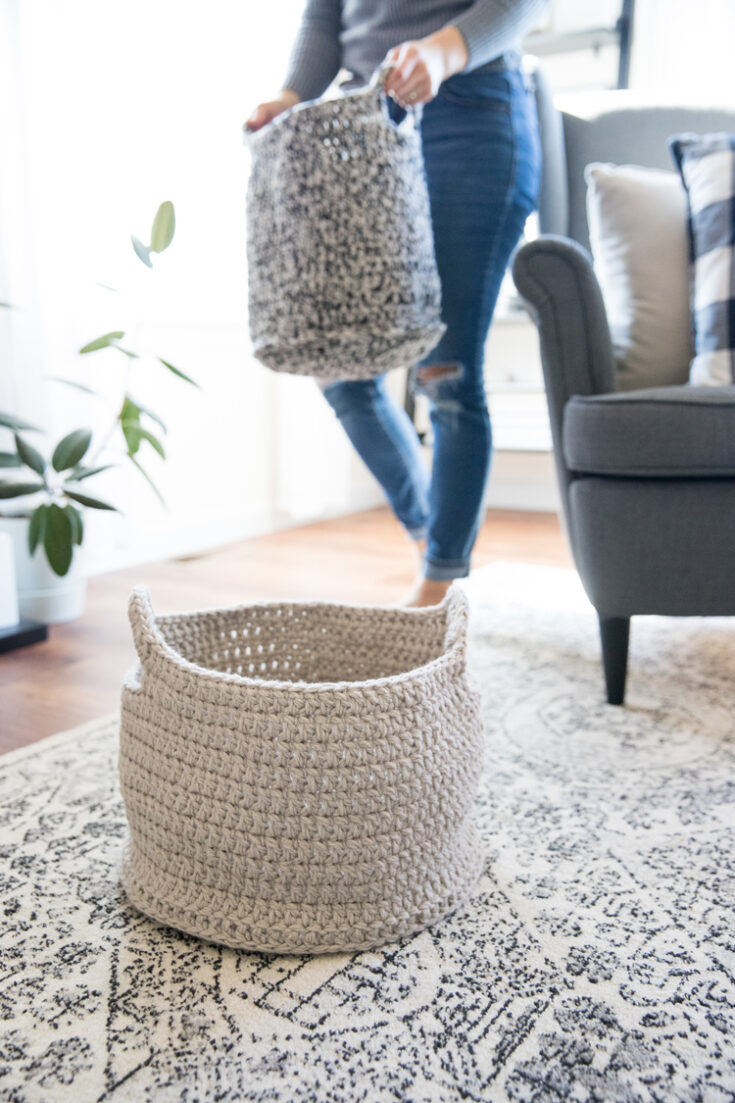 A person carries a gray woven basket while another similar basket sits on a patterned rug next to a gray armchair and a potted plant.