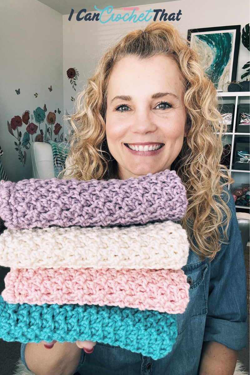 Smiling woman holding a stack of four crocheted blankets in pastel colors; shelves and floral wall art in the background.