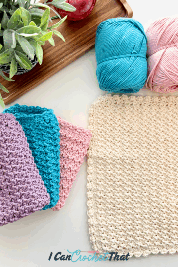 Crocheted washcloths in pastel colors, two balls of yarn, a potted plant, and a wooden tray are displayed on a white surface.
