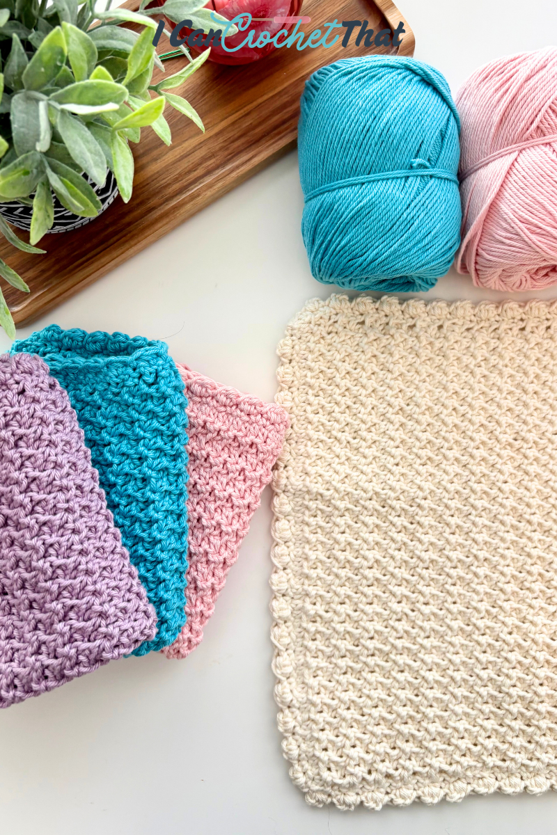 A beige crocheted square, three colorful crocheted cloths, two yarn balls, and a potted plant on a white surface.