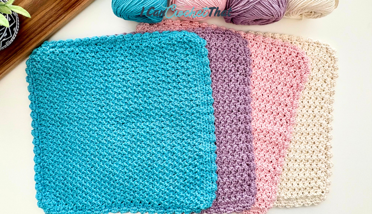 Four crocheted square dishcloths in blue, purple, pink, and beige are stacked on a white surface, with yarn balls and a plant nearby.