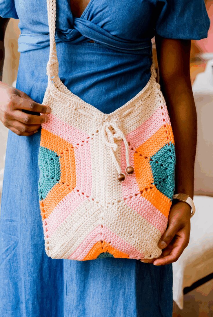 A person wearing a blue dress holds a colorful crocheted shoulder bag with geometric patterns in cream, pink, orange, and green.