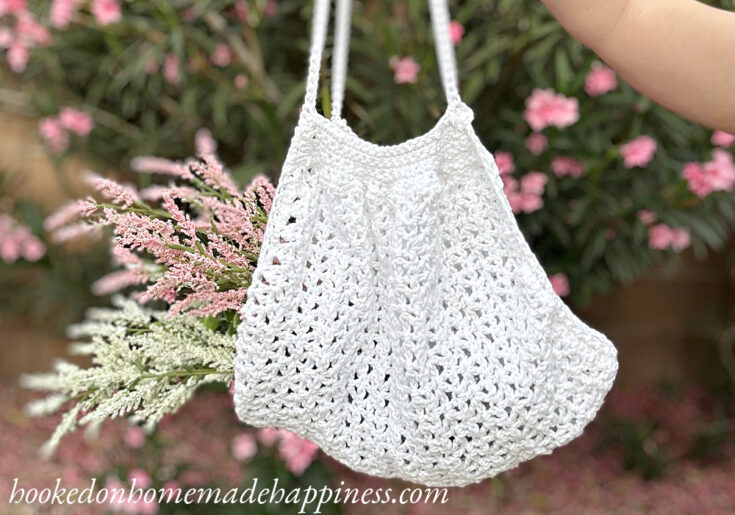 A white crocheted tote bag hangs from a hand, holding sprigs of pink and white flowers, with flowering bushes in the background.