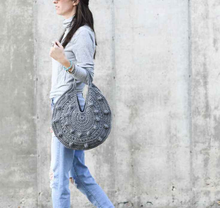 A person wearing a grey turtleneck and ripped jeans walks beside a concrete wall, carrying a large, round crocheted bag.