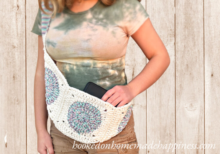 Person wearing a tie-dye shirt puts a phone into a handmade crochet crossbody bag with circular patterns, standing in front of a wooden background.