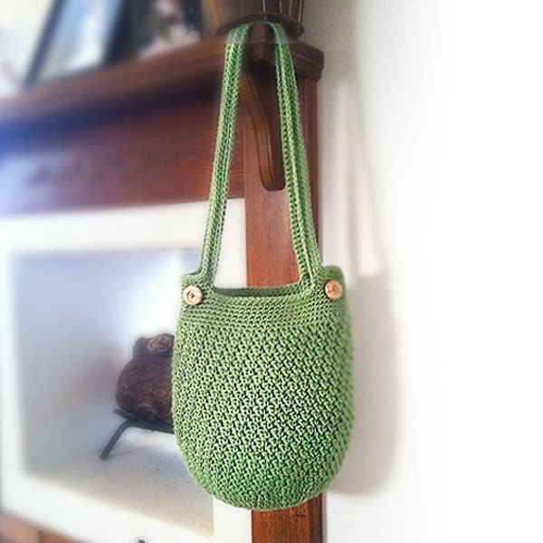 A green crocheted shoulder bag with wooden buttons hangs on a wooden hook against a white and brown wall.