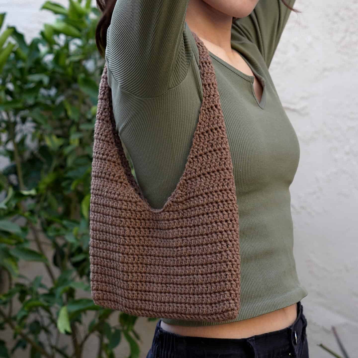 A person wearing a green long-sleeve top carries a brown crocheted shoulder bag. Green foliage and a white wall are in the background.