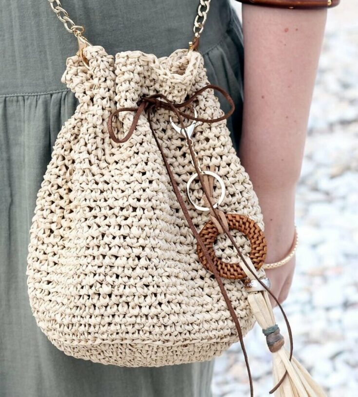 A person wearing a green dress carries a beige woven drawstring bag with decorative brown accents and a tassel keychain.