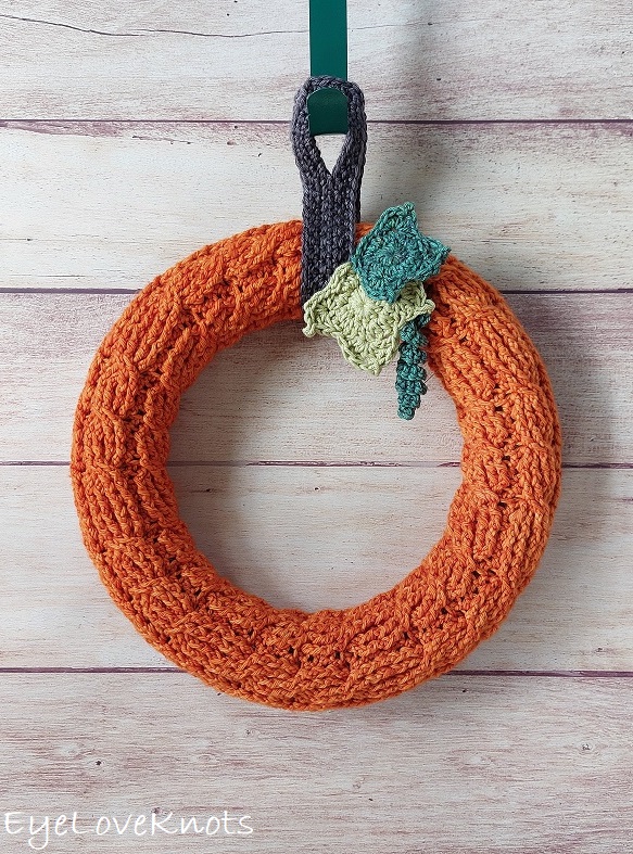 An orange crocheted wreath with textured patterns, a green and blue crocheted leaf accent, and a brown hanging loop, displayed against a wooden background.