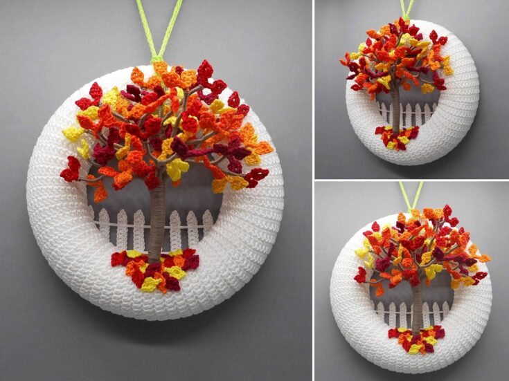 A round crocheted wreath with a white base, a small picket fence, and a tree with red, yellow, and orange leaves, hanging against a gray background.