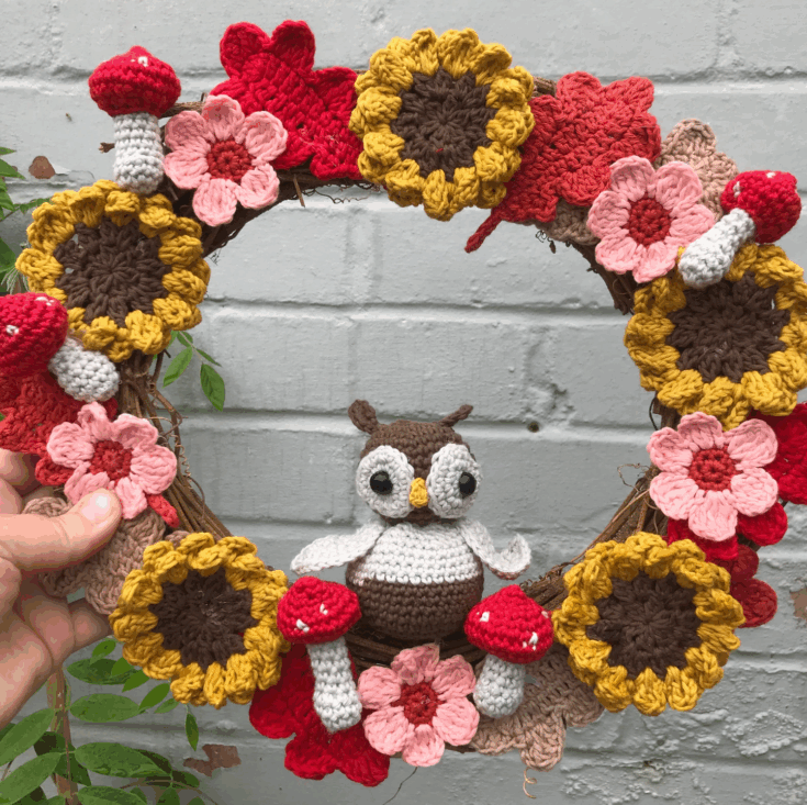 Hand holding a wreath decorated with crocheted sunflowers, mushrooms, and pink flowers, featuring a crocheted owl at the center, against a light gray brick wall.