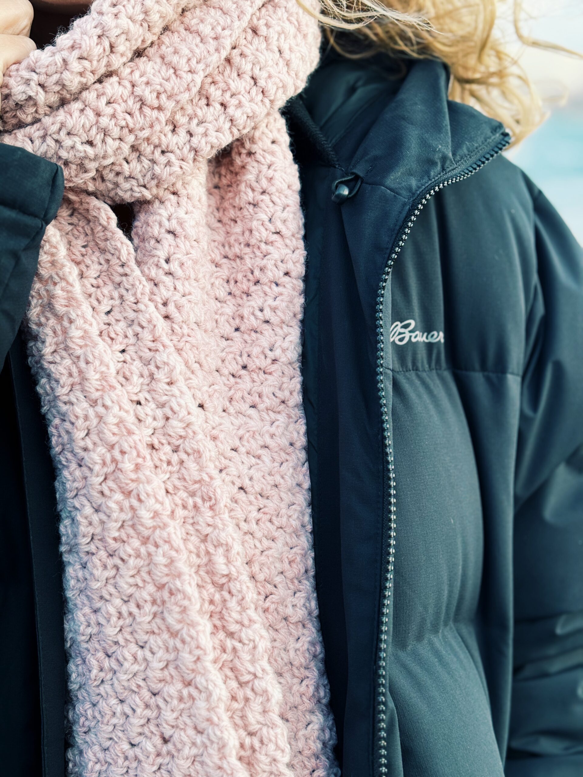 A person wearing a thick, pale pink knitted scarf over a black winter jacket.