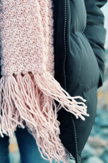 A thick, blush pink knitted scarf with fringe worn over a dark winter jacket.