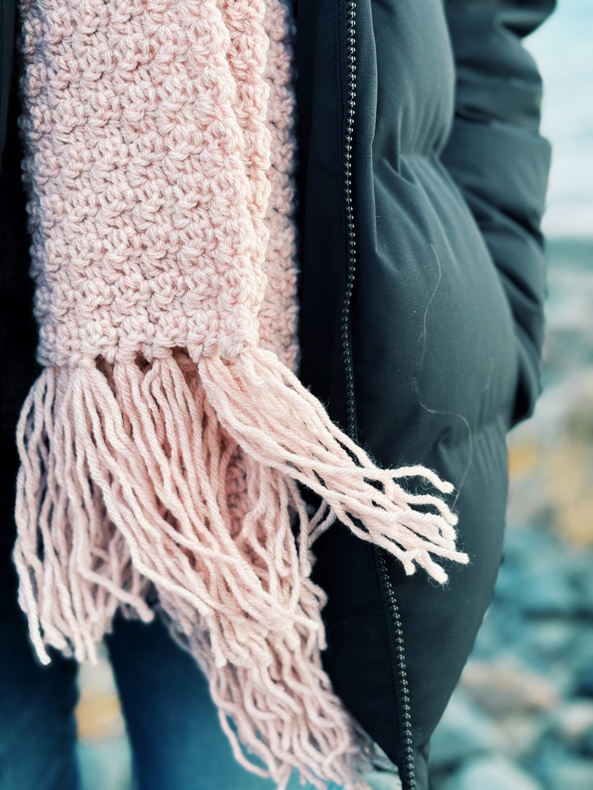 A thick, blush pink knitted scarf with fringe worn over a dark winter jacket.