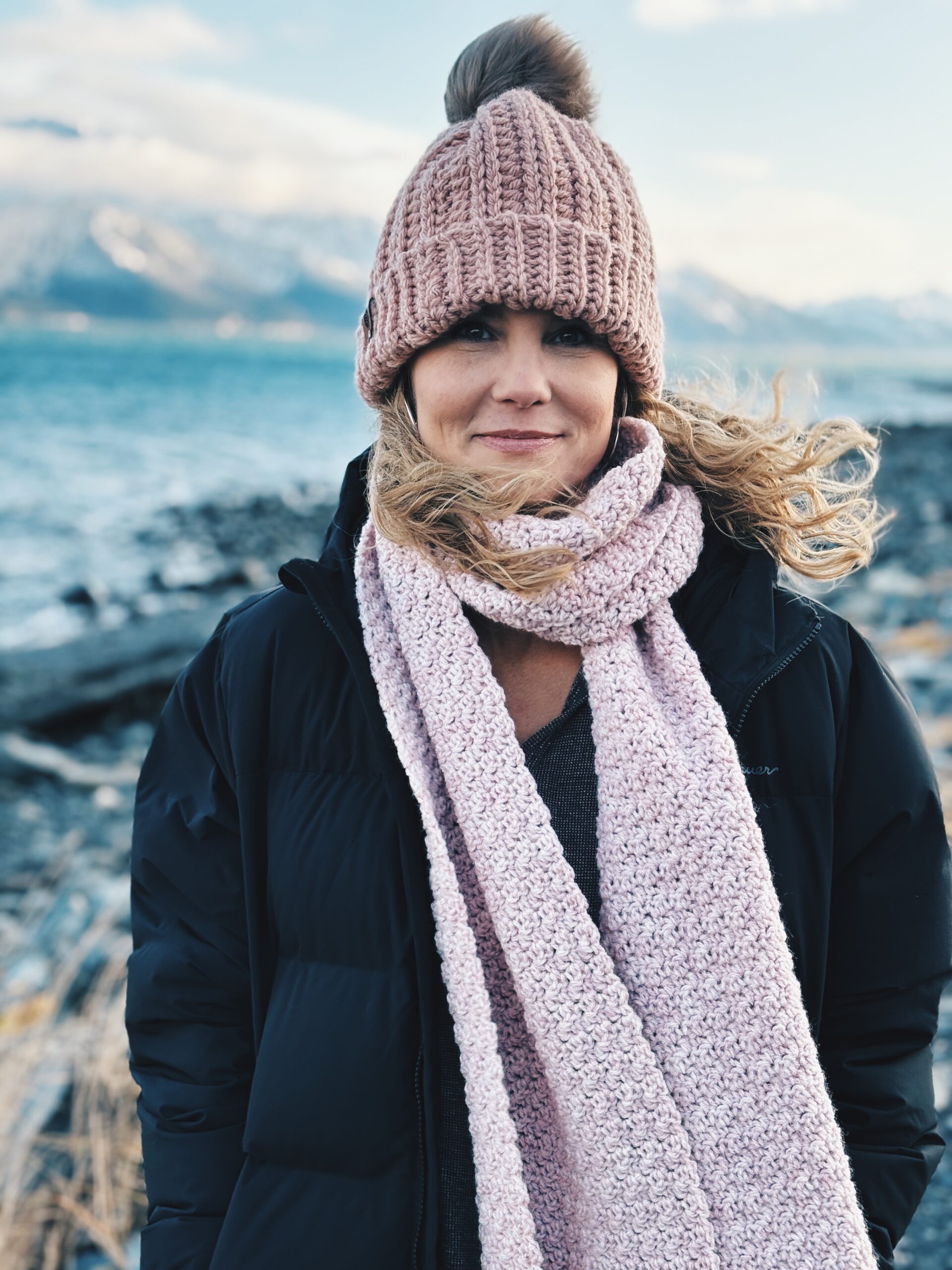 A woman wearing a pink knit hat and scarf with a black jacket, standing by a rocky shore and water with mountains in the background.