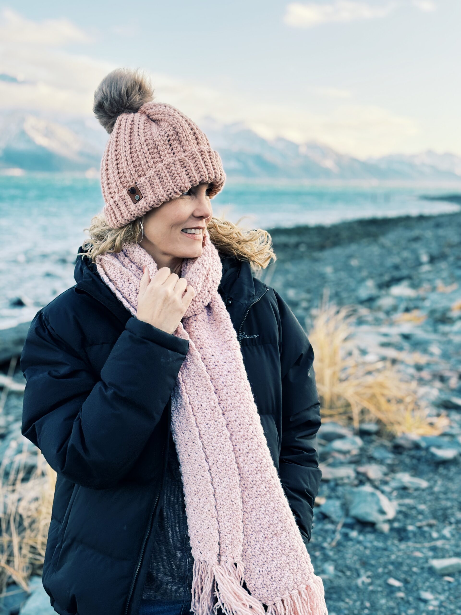 A woman wearing a black jacket, pink pom-pom beanie, and matching scarf standing on a rocky beach by a lake with mountains in the background.