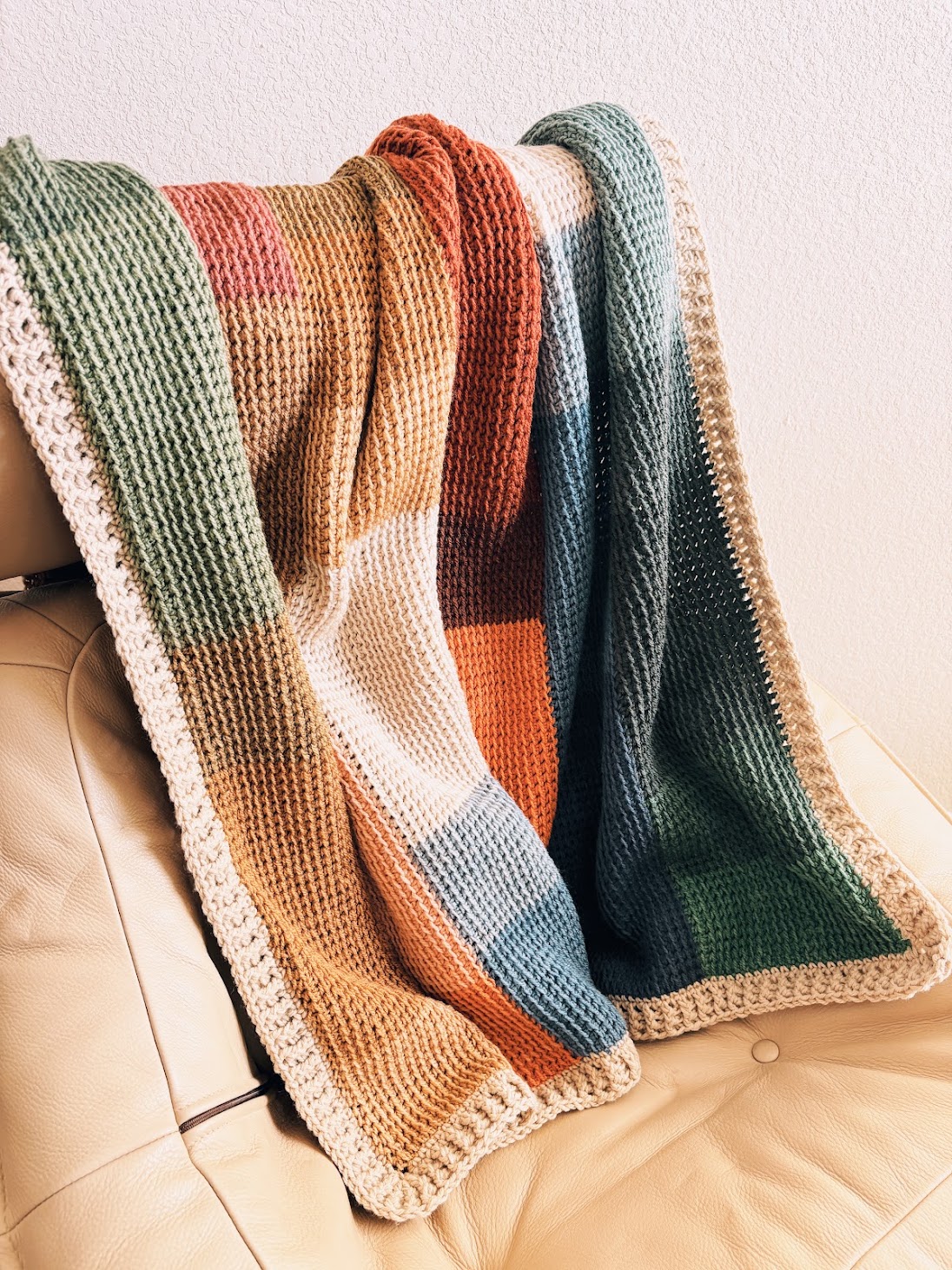 A crocheted blanket with a waffle stitch texture and a multi-colored block pattern in muted earth tones, draped over a tan leather chair.